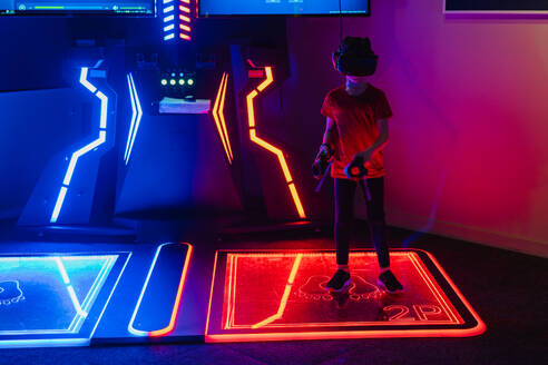 A focused player wearing a VR headset stands on a glowing red interactive gaming pad in a neon-lit arcade - ADSF53847