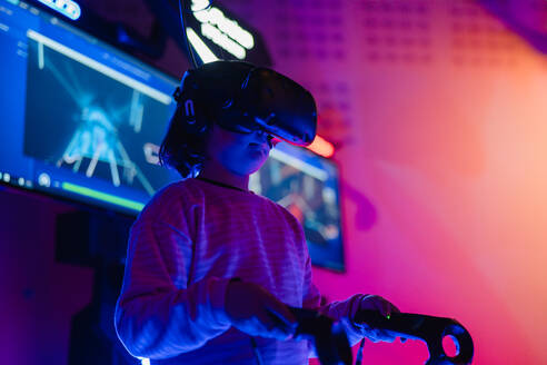 A young gamer equipped with a virtual reality headset and controllers, surrounded by vibrant neon arcade lights - ADSF53849