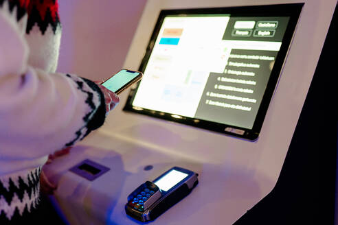 Anonymous person using a smartphone to interact with a digital kiosk touch screen display next to a card payment terminal - ADSF53851