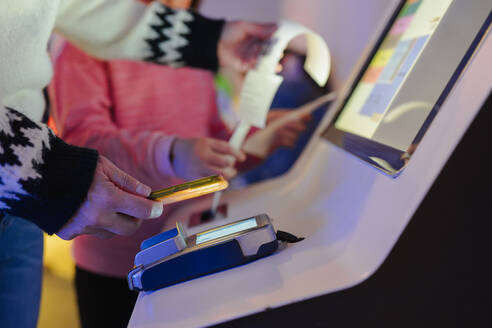 Anonymous person holding a smartphone next to a POS terminal with a receipt printing, in front of a touchscreen display - ADSF53853