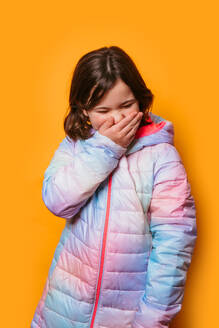 Young girl covering her mouth with her hand, wearing a colorful pastel jacket, against an orange background - ADSF53858