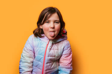 Playful girl sticking out her tongue, dressed in a multicolored jacket against a vibrant orange background - ADSF53859