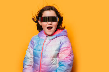 Surprised girl with mouth open wearing reflective futuristic glasses and a pastel jacket against an orange backdrop - ADSF53861