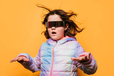 Astonished girl with windblown hair wearing futuristic glasses and a pastel coat, with arms outstretched on an orange background - ADSF53863