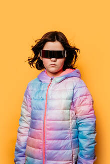 Serious young girl with windblown hair, wearing oversized futuristic glasses and a colorful puffer jacket, against a yellow background - ADSF53865
