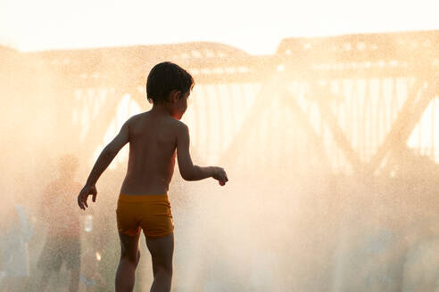 Anonymous young child enjoys a refreshing playtime in a mist of water with a cityscape backdrop as the sun sets, creating a golden hue - ADSF53878