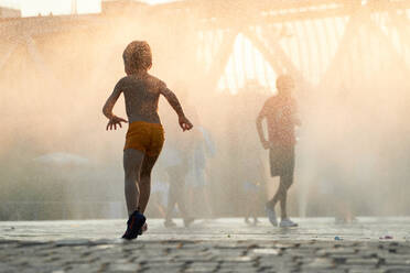 Anonymous silhouette of a child runs through the mist of a city fountain as the sun descends, casting a warm, golden glow over the urban landscape - ADSF53879