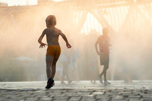 Anonymous silhouette of a child runs through the mist of a city fountain as the sun descends, casting a warm, golden glow over the urban landscape - ADSF53879