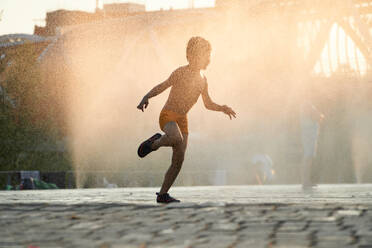 Captured in the warm glow of sunset, a joyful child dances in the water mist of an urban fountain, embodying the spirit of summer - ADSF53880