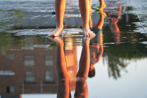 Close-up of anonymous child's feet standing by a reflective puddle, mirroring the warm glow of a city at sunset after rainfall - ADSF53881