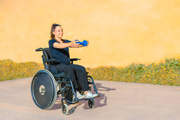 Woman in a wheelchair holding a blue kettlebell extended in front of her, engaging in an outdoor workout session - ADSF53884