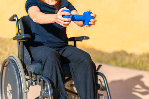 Partial view of anonymous woman in a wheelchair holding a blue dumbbell, focusing on arm exercise in a sunny outdoor setting - ADSF53885