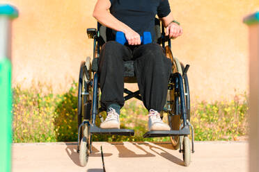 Anonymous woman seated in a wheelchair holding blue dumbbells on her lap, ready for an upper body workout session - ADSF53887