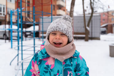 Laughing child with closed eyes in a colorful coat and woolen hat at a playground in winter - ADSF53898
