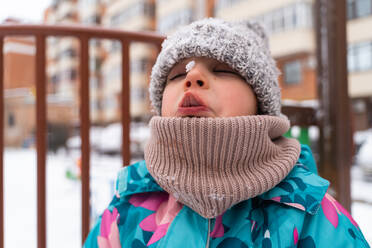 Child in a colorful jacket catching snowflakes with mouth open against a playground backdrop - ADSF53899