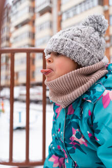 Side view of a young child with tongue out to catch snowflakes, wearing a knit hat and scarf - ADSF53900