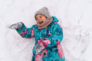 Child laughing joyfully while making a snow angel, dressed in a vibrant winter coat and hat - ADSF53901