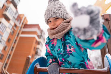 Child in a floral jacket and woolly hat playfully extending a snowball on a playground - ADSF53902