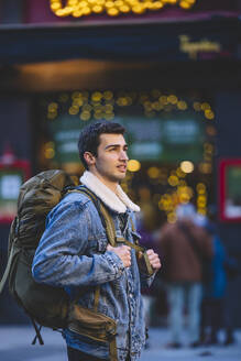 Contemplative young man with a backpack in the lively streets of Madrid, with festive lights in the background - ADSF53947