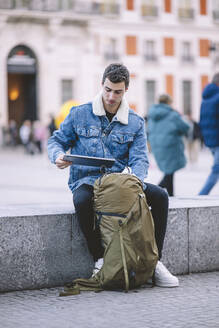 Young man with a backpack sitting on a ledge in Madrid, engrossed in his tablet - ADSF53948