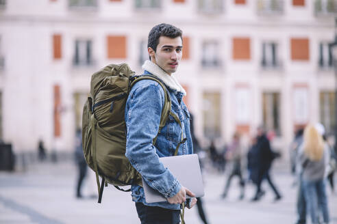 Side view of young man with backpack and laptop looking back on a busy Madrid street - ADSF53949