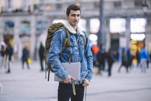 Confident young man holding a laptop stands in a bustling Madrid square looking at camera - ADSF53950