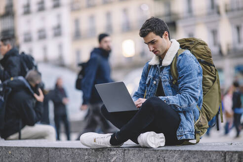 Young man works on his laptop while seated on the steps in a lively Madrid square - ADSF53951