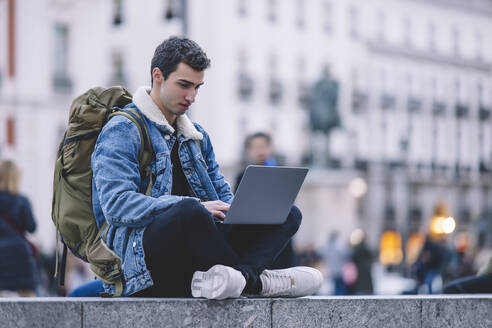 Young man works on his laptop while seated on the steps in a lively Madrid square - ADSF53952