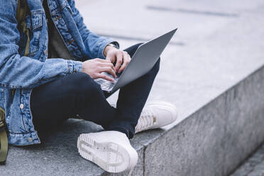 Anonymous young man works on his laptop while seated on the steps in a lively Madrid square - ADSF53953