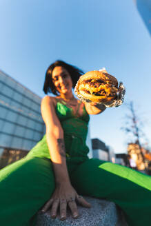 A cheerful woman in a green jumpsuit offers a juicy burger in front of the iconic Cuatro Torres business area of Madrid, Spain, showcasing a vibrant urban lifestyle. - ADSF53955