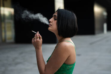 Side view of stylish young woman exhales smoke while holding a cigarette, set against the backdrop of Madrid's contemporary 4 towers area. - ADSF53961