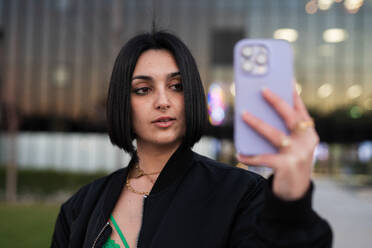 Young woman takes a selfie with a smartphone, urban style fashion, with Madrid's Four Towers in the blurry background. - ADSF53971