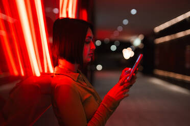 Side view of an individual engrossed in their smartphone at night, illuminated by vibrant neon lights in an urban setting. - ADSF53976