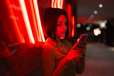 A young woman engrossed in her smartphone under vibrant neon lights, with an urban backdrop depicting the modern lifestyle in Madrid's Four Towers Business Area. - ADSF53977