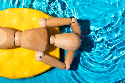 Wooden artist's mannequin relaxing on a yellow pool float in clear blue swimming pool water, simulating a summer leisure scene - ADSF54024