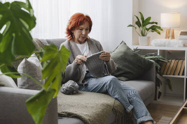 Ältere rothaarige Frau sitzt auf der Couch und strickt zu Hause - ALKF01039