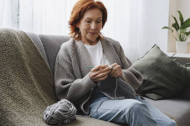 Retired redhead woman sitting on couch knitting at home - ALKF01040