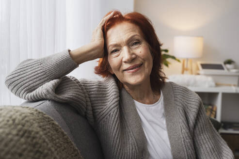 Smiling senior redhead woman sitting on couch at home - ALKF01043