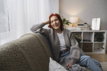 Smiling retired senior redhead woman sitting on couch at home - ALKF01044
