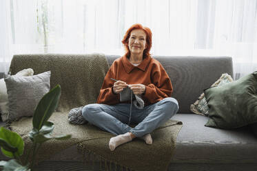 Smiling retired redhead woman sitting on couch knitting at home - ALKF01045