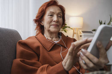 Retired senior woman using smart phone with headphones sitting on sofa in living room - ALKF01048