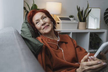 Relaxed redhead senior woman sitting with smart phone and headphones in living room - ALKF01050