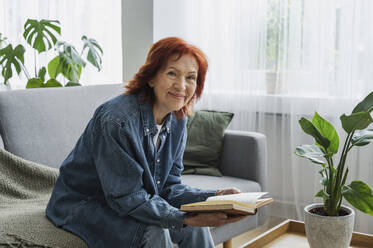 Lächelnde ältere Frau im Ruhestand liest ein Buch auf dem Sofa zu Hause - ALKF01052