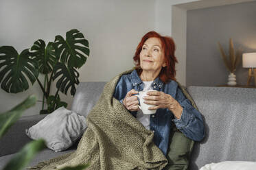 Ältere Frau sitzt entspannt unter einer Decke auf dem Sofa und trinkt zu Hause Tee - ALKF01057