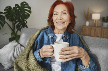 Smiling senior woman sitting under blanket on sofa drinking tea at home - ALKF01058