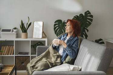 Nachdenkliche ältere Frau sitzt entspannt unter einer Decke auf dem Sofa und trinkt zu Hause Tee - ALKF01059
