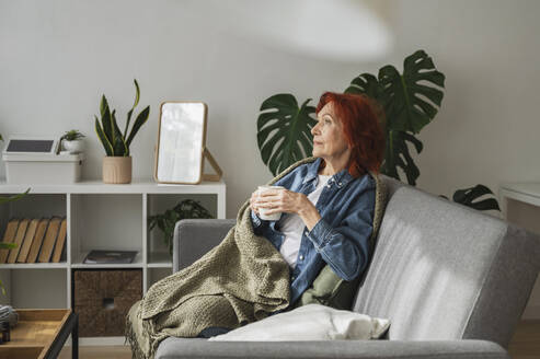 Nachdenkliche ältere Frau sitzt entspannt unter einer Decke auf dem Sofa und trinkt zu Hause Tee - ALKF01059
