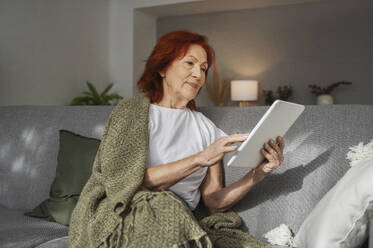 Rothaarige Rentnerin mit digitalem Tablet, die zu Hause auf dem Sofa sitzt - ALKF01064