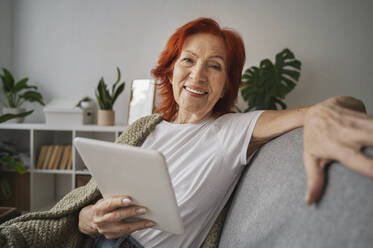 Smiling redhead senior woman with digital tablet sitting on sofa at home - ALKF01065