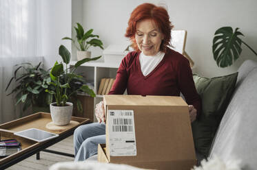 Lächelnde ältere Frau im Ruhestand öffnet Paket auf dem Sofa zu Hause - ALKF01068